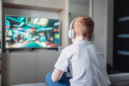 Red-haired boy in headphone sits on a couch in front of TV and playing video games