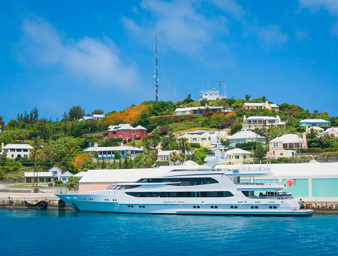A large, white,  luxury  mega yacht is docked in St. George, Bermuda with a neighborhood of colorful pastel homes in the background.