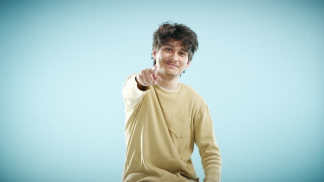 A young man pointing at you while smiling against a blue background. Portrait of an attractive guy gesturing with his index finger