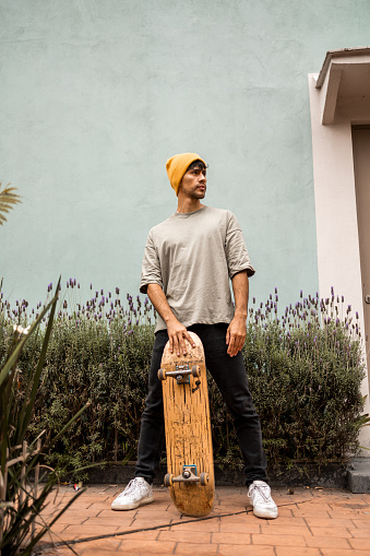 Low angle view portrait of a young Latin man wearing yellow hat looking away. He is holding his skateboard.