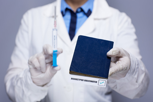 female medical doctor in white medical robe with monkeypox vaccine syringe and monkeypox vaccine passport against grey background.
