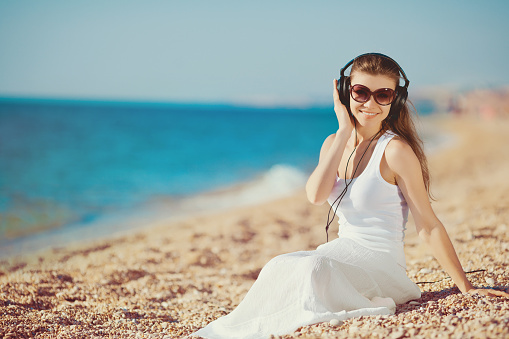 Side view of a young woman in a white dress listening to music with headphones on the beach by the sea. A woman on the beach in headphones listens to music, closed her eyes and enjoys the melody. Women's summer holidays. Relax music.