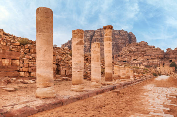 colunas de pedra vermelha permanecem na rua colonizada em petra, jordânia, montanhas rochosas com buracos de cavernas no fundo - petra ancient civilization jordan cave - fotografias e filmes do acervo