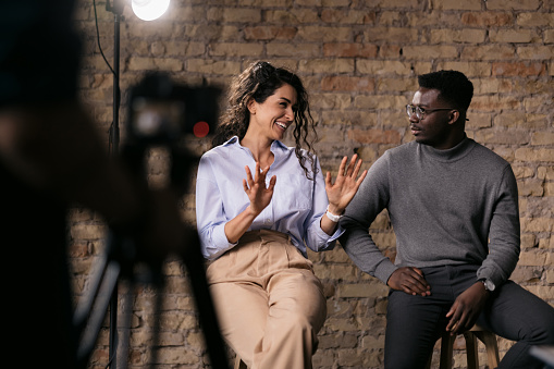 Happy diverse couple of models giving an interview in a studio, smiling and gesturing with their hands while they talk, enjoying the backstage