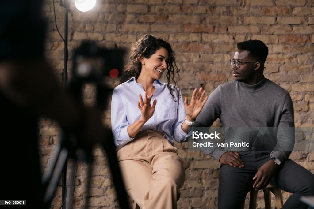 Diverse couple of models giving an interview in a studio Happy diverse couple of models giving an interview in a studio, smiling and gesturing with their hands while they talk, enjoying the backstage Home Video Camera Stock Photo