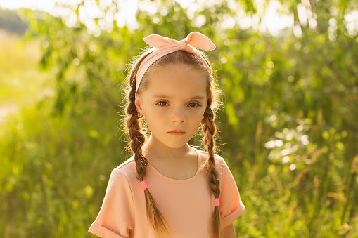 Blonde girl with long hair in pink dress against a summer background looks at the camera. Portrait.