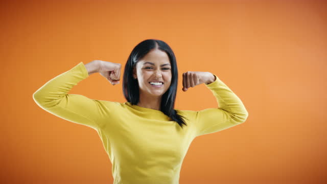 A young woman flexing her muscles against an orange studio background. Happy female smiling while raising both her arms and showing her biceps