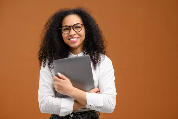 Young African American woman with afro hairstyle wearing smart casual wear and stylish eyeglasses standing isolated on brown , carrying laptop computer, smiling, female office employee stock photo