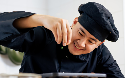 Portrait, close up handsome Asian Japanese professional chef wearing black uniform, smiling with happiness, cooking and decorating dish. Hotel or Restaurant Service Concept