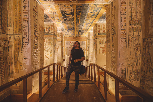 Low angle full length faceless stylish female traveler sitting on stone construction and admiring historic architecture in ancient ruined heritage building with hieroglyphs on weathered columns. Travel concept