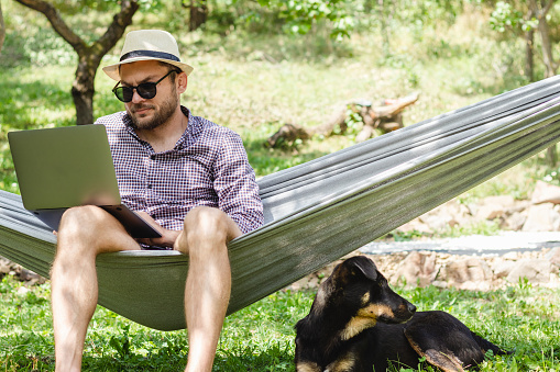 Man in hat lying down in hammock and using laptop, his dog lying down under hammock near the owner.