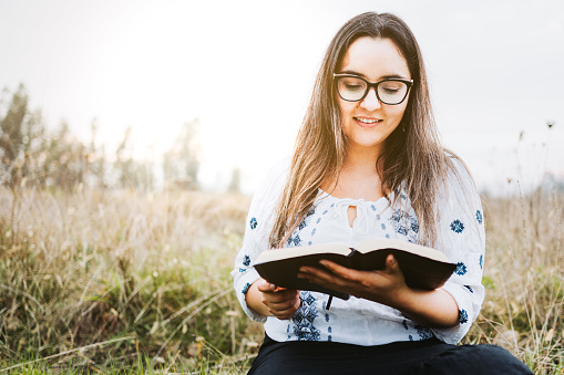 woman with glasses sitting on the grass holding and reading an open Bible. Selective focus. Horizontal. High quality photo. Copy space