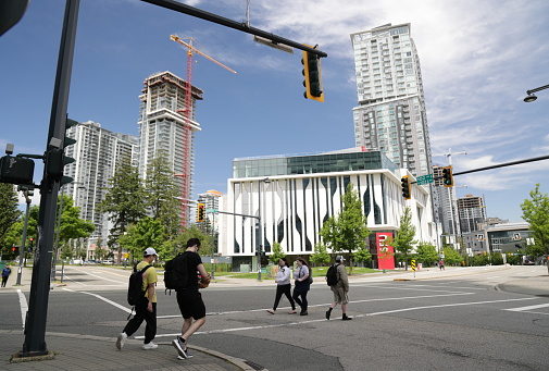 Surrey, Canada - June 17, 2022: Pedestrians cross 102nd Avenue in Surrey City Centre. Background shows the Simon Fraser University Surrey on University Drive (SRYE). Residential buildings under construction line the downtown district.