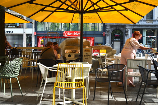 Antwerp, Belgium - June 18, 2022: caucasian hetero couple having breakfast on a cafe terrace under a big beautiful yellow parasol named Panos, company that sells croissants and bread.