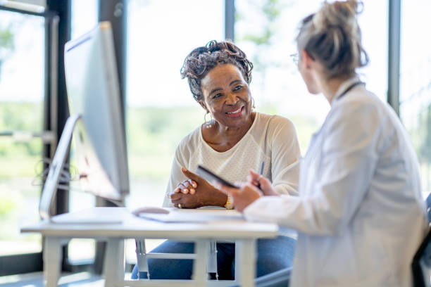 African Woman at a Medical Appointment A senior woman of African decent, sits with her young doctor at her desk as they meet to discuss her health. The woman is dressed casually and smiling at the doctor as she listens attentively. The doctor is holding a tablet as she takes notes electronically for the woman's file. old woman smiling tablet stock pictures, royalty-free photos & images
