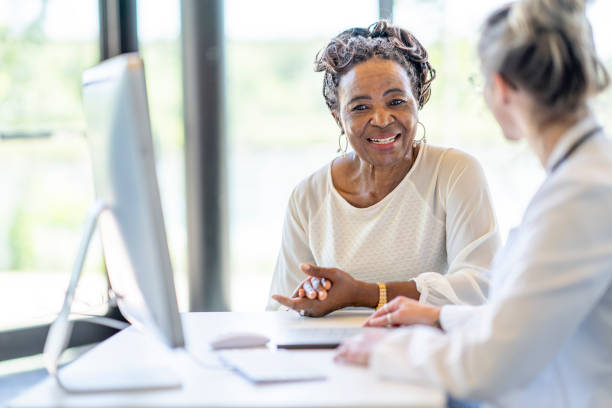 African Woman at a Medical Appointment A senior woman of African decent, sits with her young doctor at her desk as they meet to discuss her health. The woman is dressed casually and smiling at the doctor as she listens attentively. The doctor is holding a tablet as she takes notes electronically for the woman's file. old woman smiling tablet stock pictures, royalty-free photos & images