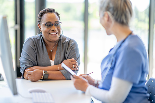 A middle aged woman of African decent, sits with her doctor at her desk as they meet to discuss her health.  The woman is dressed casually and smiling at the doctor as she listens attentively.