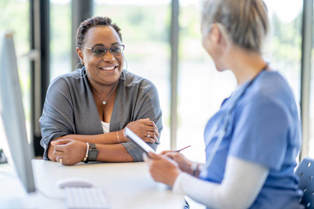 African Woman at a Medical Appointment A middle aged woman of African decent, sits with her doctor at her desk as they meet to discuss her health.  The woman is dressed casually and smiling at the doctor as she listens attentively. doctor consultation stock pictures, royalty-free photos & images