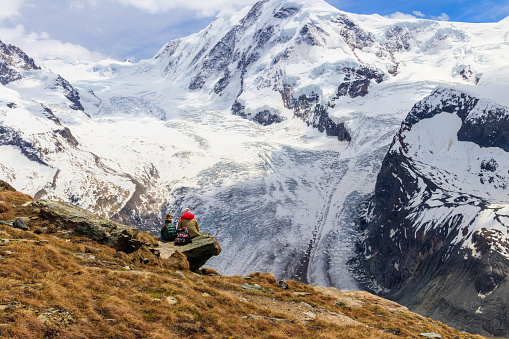 Mother and daughter sitting on a stone and looking at panorama of the Pennine Alps with famous Gorner Glacier and impressive snow capped Monte Rosa Massif close to Zermatt, Switzerland