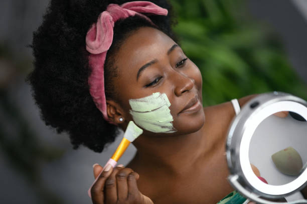Beautiful young woman applying facial mask. Beautiful young woman applying facial mask. She's sitting at the table, looking at mirror and taking care of here skin. centella asiatica cream stock pictures, royalty-free photos & images