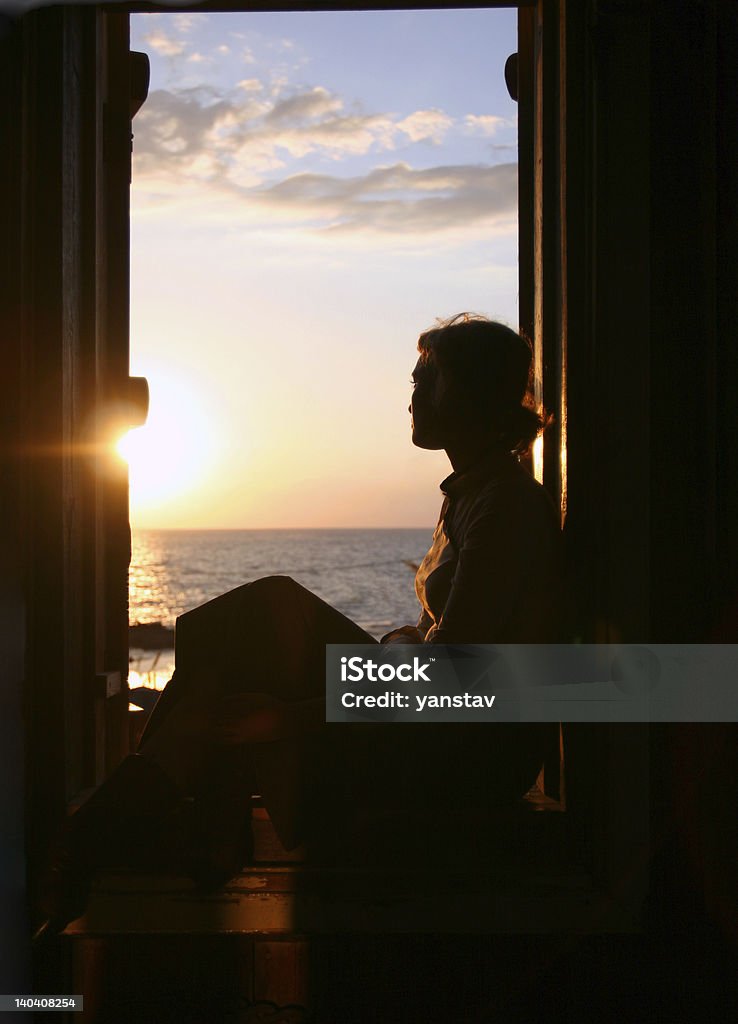 girl looks at the sea. A silhouette of the young woman sitting in a window on a background of a sea decline.The photo is made in the evening, in a small old house on coast of the mediterranean sea. The sun slowly fell to this silent evening for horizon, gently concerning with the warm, kind beams of the person of the girl dreaming of close happiness Window Stock Photo
