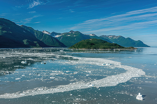 Disenchantment Bay, Alaska, USA - July 21, 2011: Floating ice circle on blue water under blue cloudscape with forested and snow covered mountain range leading to ocean.