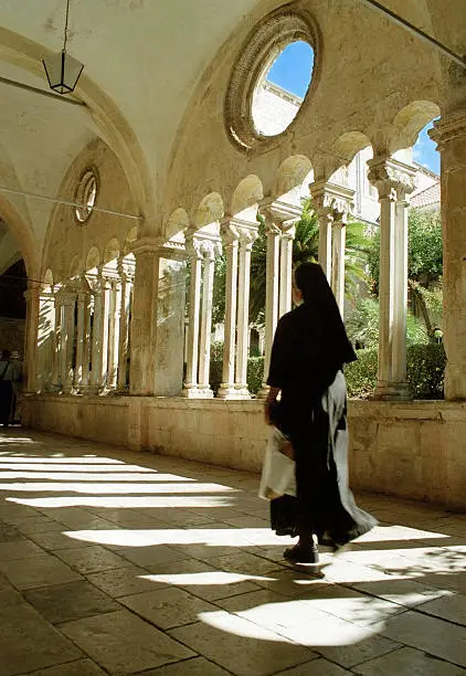 Nun walking along an arched corridor by the internal courtyard of the Franciscan Monastery, Dubrovnik, Croatia.