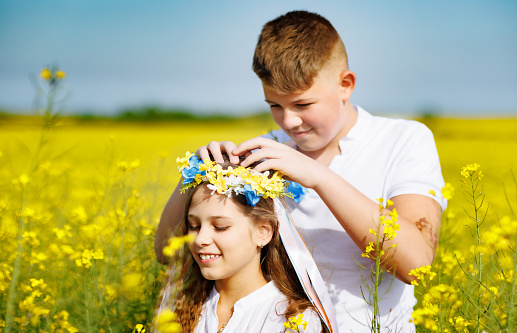 Cheerful laughing teenage guy throws up multi-colored long ribbons in bright Ukrainian flower wreath on at his cute ruddy smiling sister on her head, against clear sky and blooming yellow fields