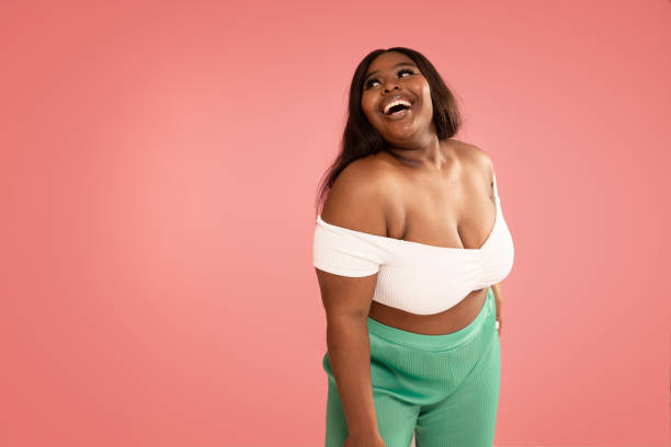 Joyful Plus Size african Woman laughing, posing on a pastel pink Studio Background. Real people emotions, toothy smile. Fashionable young girl in white top and green pants. Body positive. Joyful Plus Size african Woman laughing, posing on a pastel pink Studio Background. Real people emotions, toothy smile. Fashionable young girl in white top and green pants. Body positive. A lot of copy space. plus size woman fashion laughing stock pictures, royalty-free photos & images