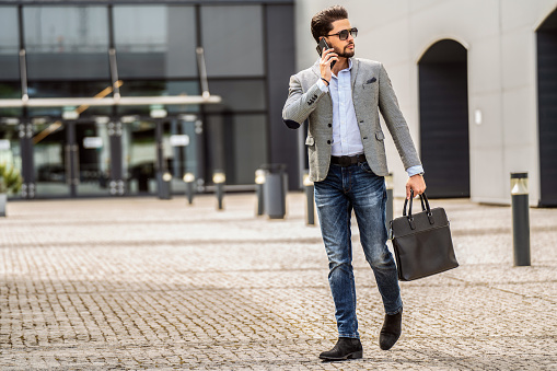 Handsome businessman walking on the street, talking via mobile phone. Fashionable style. Male model. Outdoor photo.