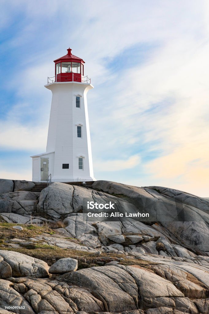 Lighthouse Sitting on Rocks with Clouds in the Sky Peggy’s cove lighthouse in Nova Scotia sitting on top of rocks with sky behind. Blue and orange sky with clouds Lighthouse Stock Photo