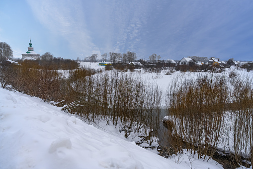 Winter panoramic view of the valley of an unfrozen river with snow-covered banks, trees, an old church and wooden houses of the townspeople. Solikamsk (Ural, Russia). Blue sky with cirrus clouds.