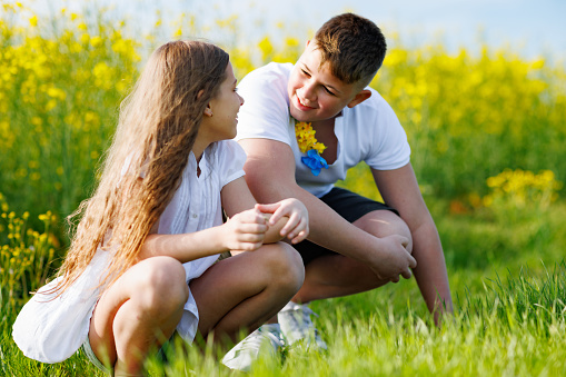 Native joyful teenage children enjoying warm weather: big strong kind brother and little smiling dreamy sister sit on squats and talk in field with yellow flowers and green grass under blue sky