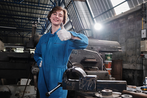 One cheerful young Caucasian female industry worker in safety uniform relaxes and thumbs up after work success and expresses smile happiness in a mechanical factory. Professional engineer occupation.