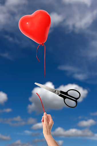 Scissors cutting the string of a hand holding heart-shaped balloon against blue sky.