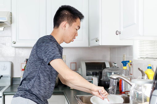 A teenage boy is in the kitchen washing dishes. He is focusing at the sink. The photograph is a side view shot.