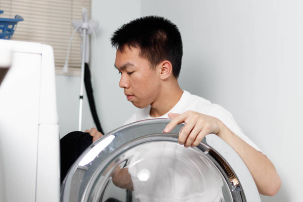 An Asian teenage boy is doing the laundry at home with a laundry machine. He is holding a shirt.