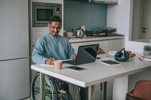 Asian indian man in wheelchair sitting at dining room e-learning with laptop and writing notes