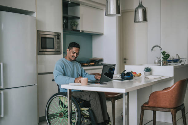 istock Asian indian man in wheelchair sitting at dining room e-learning with laptop and writing notes 1404119805