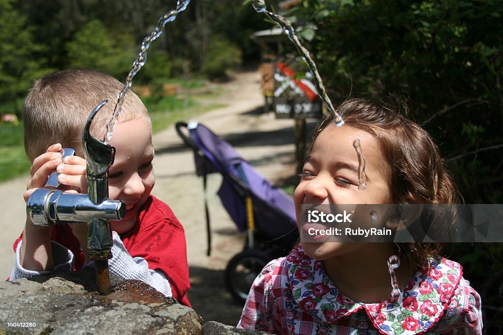 SWEET LAUGHTER Laughing together, a little boy turns on a water fountain for a little girl, while she tries to taste the overflowing stream of water. Drinking Water Stock Photo