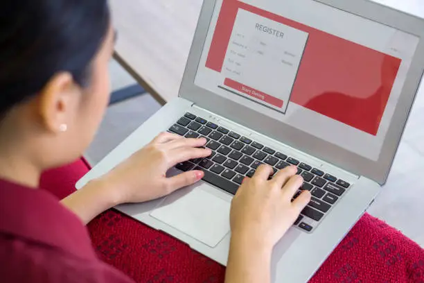 Photo of Young asian woman in red dress typing on laptop computer keyboard, registering for online dating application on website.