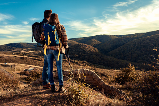 Rear view of couple with backpacks hiking together on countryside during sunset.