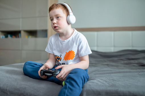 Boy in headphones with a gamepad playing video games sitting on a bed in bedroom, boredom emotions