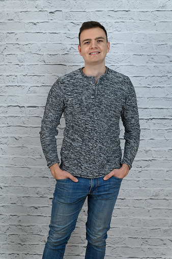 Young attractive man in a gray jumper and blue jeans. A man stands against the background of a light brick wall.