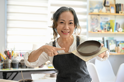 Professional aged Asian female potter showing her ceramic plate in her studio. Beautiful aged Asian woman joining a pottery workshop.