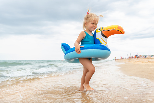 Portrait of cute little caucasian blond toddler baby girl in swimsuit and toucan inflatable ring toy enjoy swimming on sand sunny seaside beach on bright summer day. Family holiday vacation fun.
