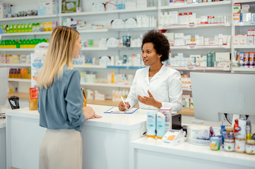 Pharmacy Drugstore: Beautiful Female Cashier in White Coat Serves Customer. Medicine Recommendation, Advice, Talking. Drugstore With Full of Drugs, Pills, Health Care