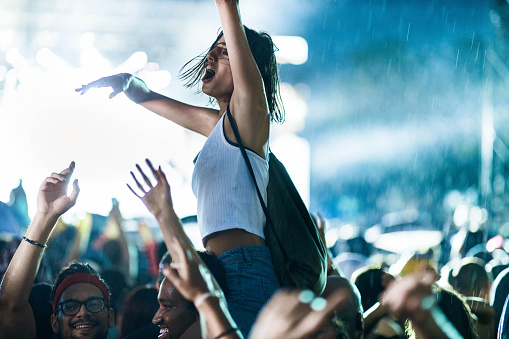 Closeup rear view of large group of people enjoying an open air concert on a summer night. There are a lot of hands raised. Blurry stage lights in background