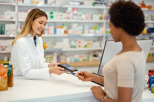 Pharmacy Drugstore Checkout Cashier Counter: Professional Female Pharmacist Sells Medicine, Black Female Customer Pays Using Contactless Payment Terminal and Credit Card