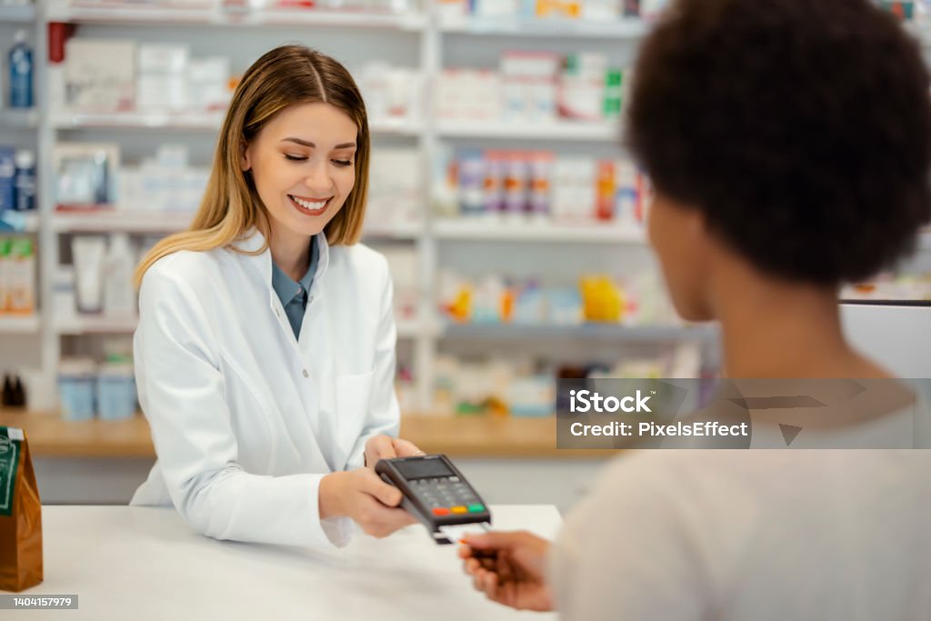 Accepting payment for a healthcare purchase Pharmacy Drugstore Checkout Cashier Counter: Professional Female Pharmacist Sells Medicine, Black Female Customer Pays Using Contactless Payment Terminal and Credit Card Paying Stock Photo